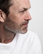 Side profile of a mature man with a beard wearing a white T-shirt against a gray background.